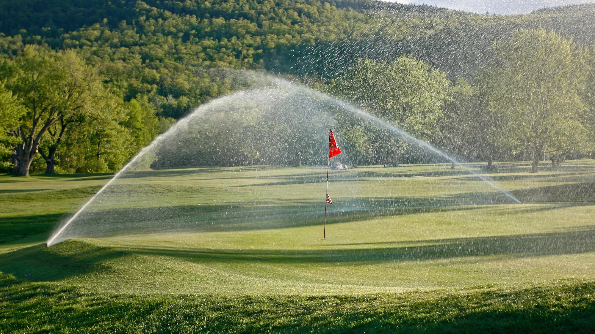 Sprinkler arcing water across a golf green with the pin still in, wooded hills behind